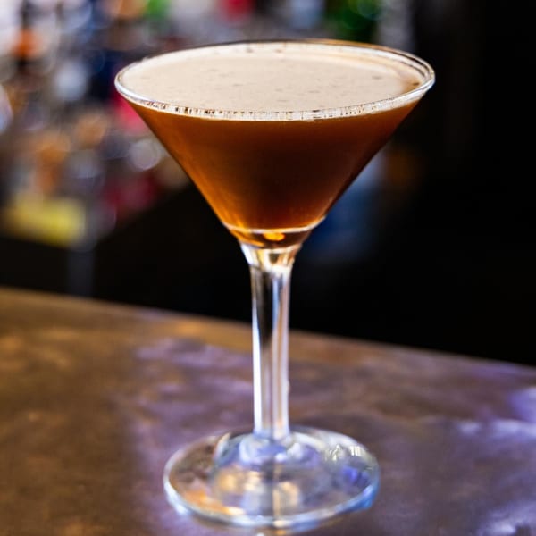 A dark brown espresso martini with a foamy top in a clear martini glass on a bar.