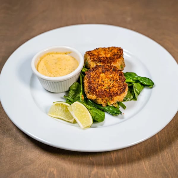 Two golden-brown crab cakes on a bed of spinach with a side of sauce and lime wedges.